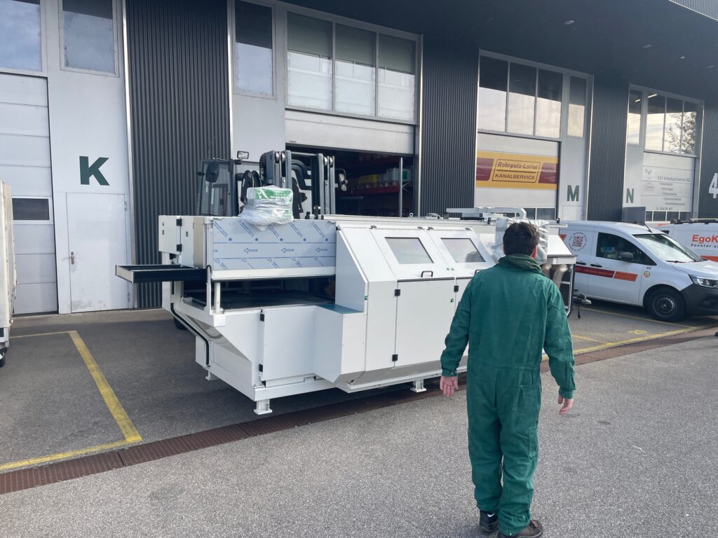 Ein Arbeiter in einem grünen Overall geht auf eine große, weiße Industriemaschine vor einem Lagerhaus zu, in dessen Nähe Fahrzeuge geparkt sind.