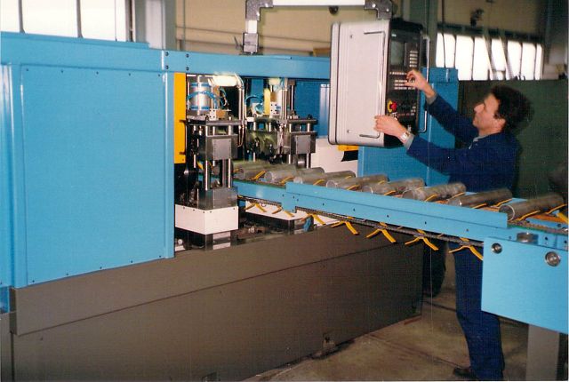 Ein Arbeiter bedient eine große blaue Maschine mit einem Bedienfeld und überwacht ein Fördersystem, durch das Produkte transportiert werden.